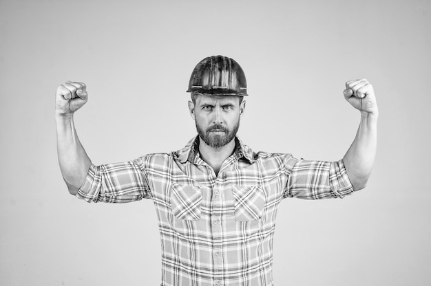 Gutaussehender Baumeister mit Bauschutzhelm und kariertem Hemd demonstriert Macht auf der Baustelle männliche Stärke