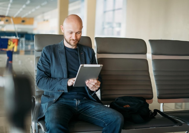 Gutaussehender erwachsener kahlbärtiger Geschäftsmann im Anzug mit Tablet in der Flughafenlounge