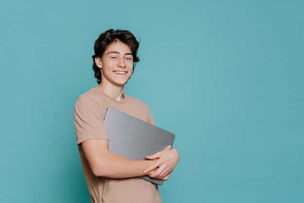 Gutaussehender italienischer fröhlicher Student, der einen Laptop trägt, blickt auf die Kamera, breites Lächeln steht vor türkisfarbenem Hintergrund mit Kopierplatz für Werbung Mockup Jugend- und Studentenleben Junge mit neuem Computer