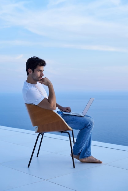 Gutaussehender junger Mann, der sich zu Hause auf dem Balkon des Laptops entspannt und arbeitet, während er den Sonnenuntergang betrachtet