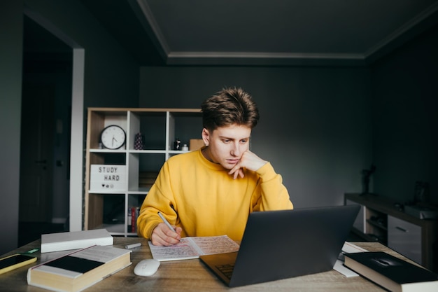 Gutaussehender junger Mann, der zu Hause Hausaufgaben auf dem Desktop vor dem Hintergrund des Zimmers macht, auf den Laptop-Bildschirm schaut und in ein Notizbuch schreibt