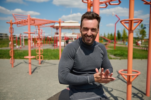 Gutaussehender kaukasischer Mann, Macho, Sportler, Athlet, lächelnd mit zahnigem Lächeln, Blick in die Kamera auf dem Sportplatzhintergrund. Glücklicher attraktiver Kerl, der den Rest nach dem Training genießt