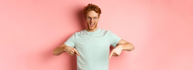 Foto gutaussehender kaukasischer mann mit rotem, unordentlichem haar und brille, der mit dem finger zeigt und auf die promo von herabblickt