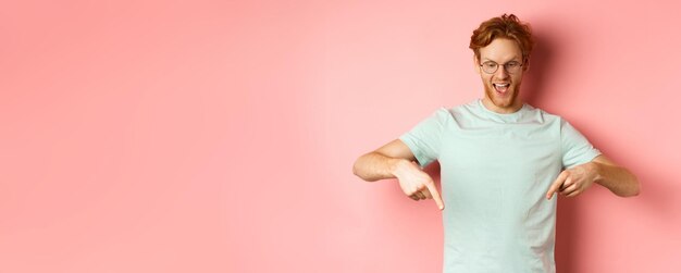 Foto gutaussehender kaukasischer mann mit rotem, unordentlichem haar und brille, der mit dem finger zeigt und auf die promo von herabblickt