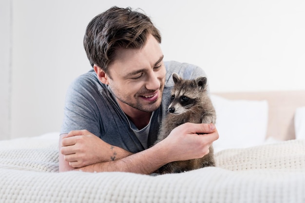 Gutaussehender lächelnder Mann, der sich mit entzückendem Waschbär auf Bettzeug ausruht