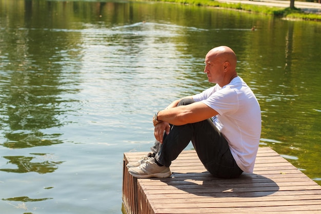 Gutaussehender Mann am See an einem sonnigen, ruhigen Tag, der auf einem Holzsteg sitzt und denkt oder
