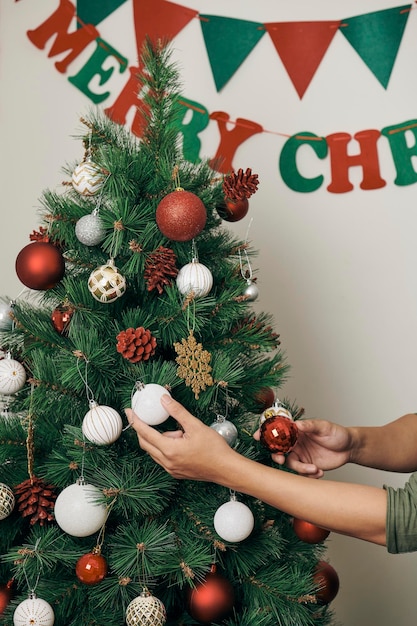 Gutaussehender Mann, der den Weihnachtsbaum mit Kugeln schmückt und zu Hause in die Kamera blickt