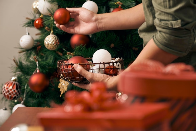 Gutaussehender Mann, der den Weihnachtsbaum mit Kugeln schmückt und zu Hause in die Kamera blickt