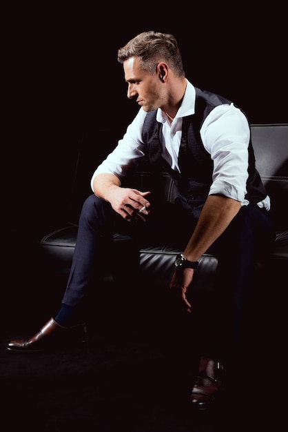 Gutaussehender Mann in formeller Kleidung sitzt auf der Couch mit Zigarette isoliert auf schwarz