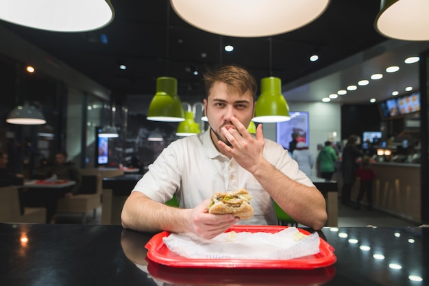 gutaussehender Mann isst einen Burger in einem Fast Food und leckt sich die Finger. Fast Food ist ein Konzept.