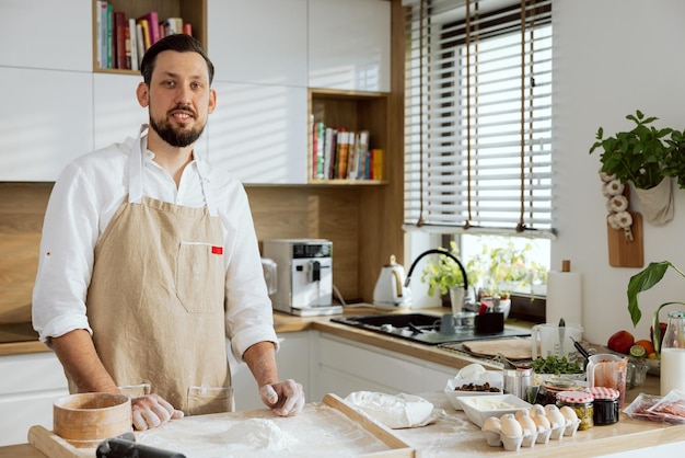 Gutaussehender Mann mit Schürze, der am Tisch steht und Mehl auf einer Holzoberfläche siebend, um Pizza zuzubereiten