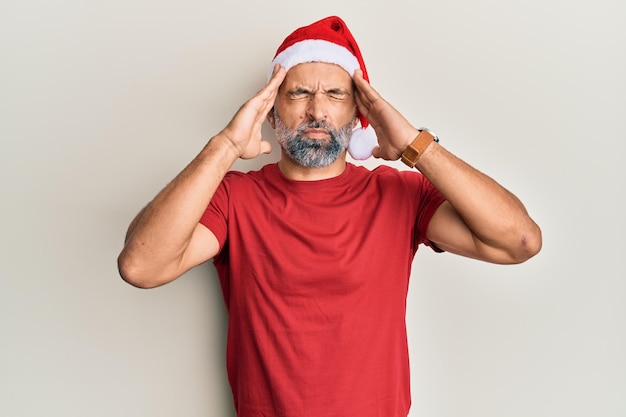 Gutaussehender Mann mittleren Alters mit Weihnachtsmütze und Sommer-T-Shirt mit der Hand auf dem Kopf, Kopfschmerzen wegen Stress und Migräne