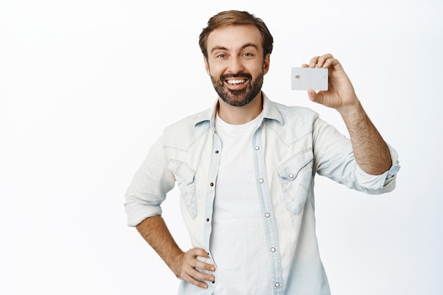Gutaussehender moderner Mann mit Bart, der Kreditkarte zeigt und zufrieden lächelnd den weißen Hintergrund des Produkts empfiehlt
