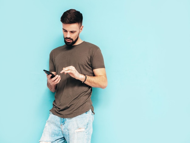 Gutaussehender, selbstbewusster, stilvoller Hipster-Lambersexual-ModelMann gekleidet in T-Shirt und Jeans Modemann isoliert auf blauer Wand im StudioHolding-Smartphone Blick auf Handy-Bildschirm Verwendung von Apps