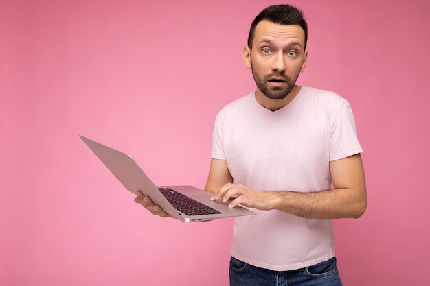 Gutaussehender überraschter und erstaunter Mann, der einen Laptop-Computer hält, der die Kamera anschaut und im T-Shirt eine SMS schreibt