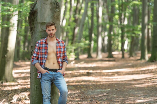 Gutaussehender unrasierter Mann mit lumbersexuellem Look im aufgeknöpften Holzfällerhemd-Waldhintergrund