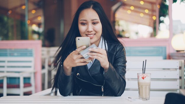 Foto gutaussehendes asiatisches mädchen tätigt online-zahlungen mit kreditkarte und smartphone, sitzt am tisch in einem open-air-café und lächelt einkaufs- und technologiekonzept