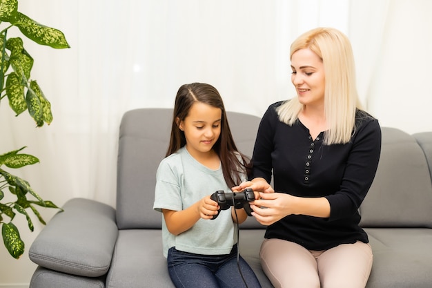 Gute Beziehung süßes kleines Mädchen mit junger Mutter mit Joystick beim Videospiel, das zusammen im Wohnzimmer sitzt und den Familienurlaub genießt.