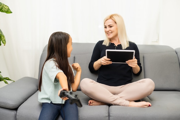 Gute Beziehung süßes kleines Mädchen mit junger Mutter mit Joystick beim Videospiel, das zusammen im Wohnzimmer sitzt und den Familienurlaub genießt.