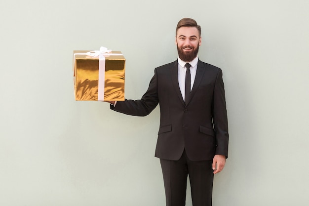 Gute Emotionen. Chef, der eine goldene Geschenkbox und ein zahniges Lächeln hält. Studioaufnahme. Grauer Hintergrund