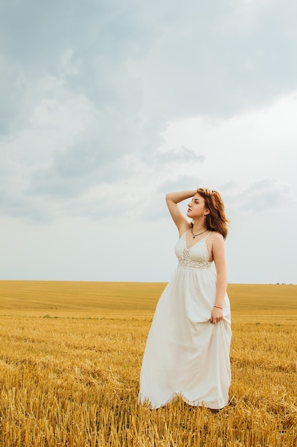 Gute Erholung im Freien. Junge schöne rothaarige Frau in der Mitte eines Weizenfeldes, Spaß habend. Sommerlandschaft, gutes Wetter. Windiger Tag mit Sonne und Wolken. Weißes Baumwollkleid im Öko-Stil.