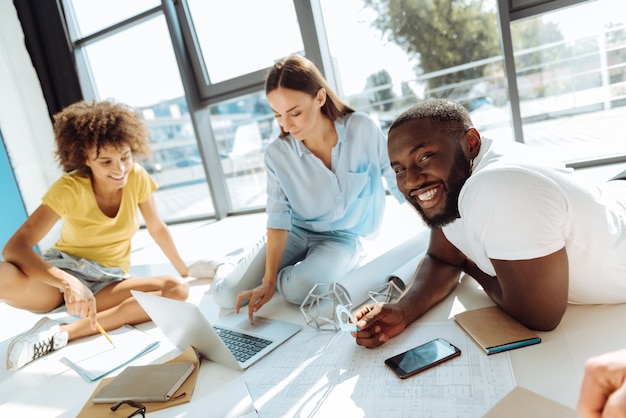 Gute Ideen. Positiv lächelnde internationale Studenten, die auf dem Boden sitzen und studieren, während sie gemeinsam am Universitätsprojekt arbeiten