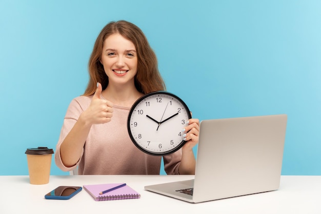 Gute Jobzeit zur Arbeit Positive glückliche Frau zufrieden Büroangestellte sitzt am Arbeitsplatz mit großer Uhr und zeigt Daumen nach oben wie Geste Indoor-Studioaufnahme isoliert auf blauem Hintergrund