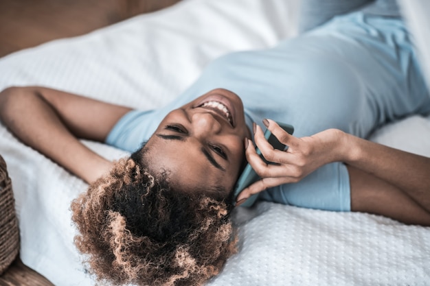 Gute Laune. Fröhliche Afroamerikanerfrau im hellen T-Shirt, das auf Smartphone kommuniziert, während zu Hause auf Bett liegend