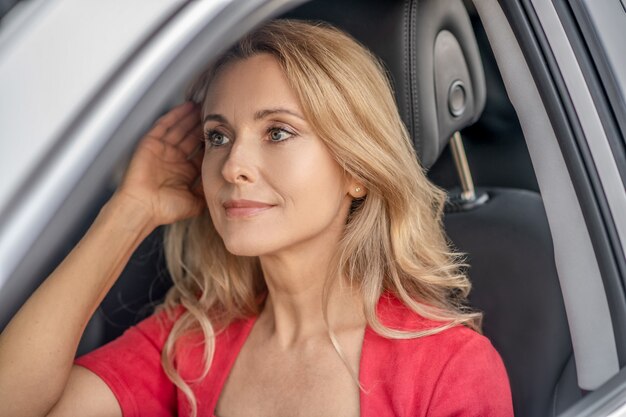 Gute Laune. Hübsche Frau in einem roten Kleid, das in einem Auto sitzt und positiv schaut