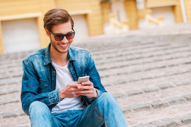 Gute Zeit in seiner Stadt verbringen. Selbstbewusster junger Mann, der beim Sitzen im Freien Handy hält