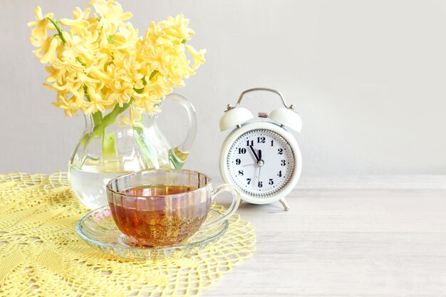 Guten Frühlingstag ein Strauß gelber Hyazinthen ein weißer Wecker eine Tasse Tee auf einer gelben durchbrochenen Serviette auf grauem Hintergrund Nahaufnahme Platz für Text