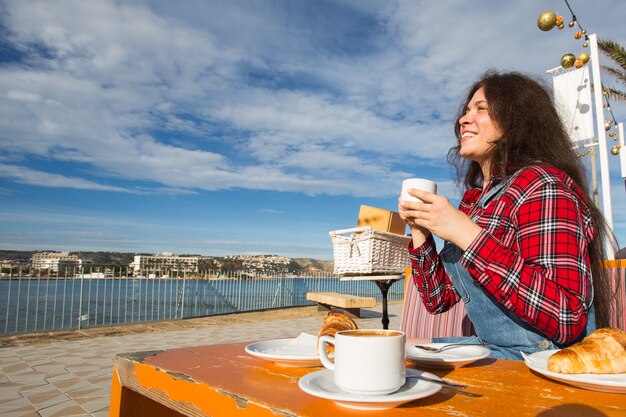 Guten Morgen. Junge Frau, die ein französisches Frühstück mit Kaffee und Croissant, die draußen an der Caféterrasse am Meer sitzen.