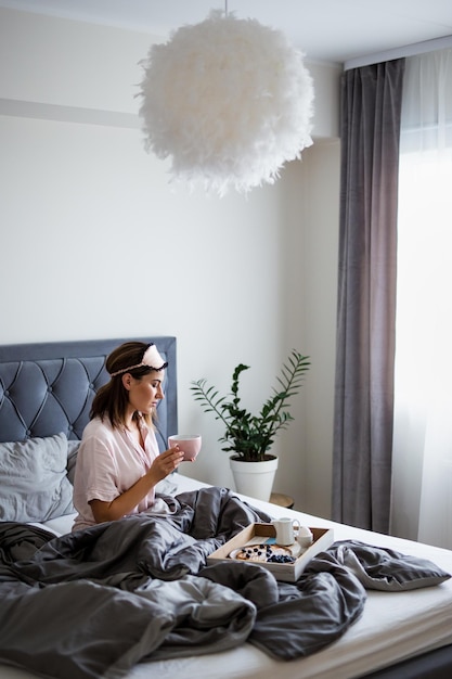 Guten Morgen Konzept - Porträt einer jungen schönen Frau im Schlafanzug, die auf dem Bett sitzt und im Schlafzimmer Kaffee trinkt