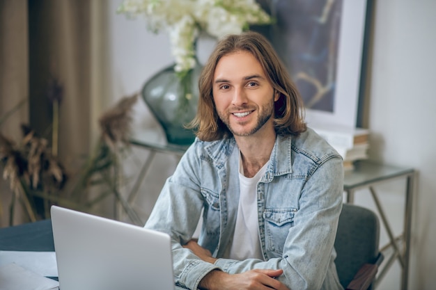 Gutes Ergebnis. Langhaariger Mann im Jeanshemd, der am Laptop arbeitet und glücklich schaut