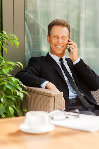 Gutes Geschäftsgespräch. Fröhlicher reifer Mann in formeller Kleidung, der mit dem Handy spricht und lächelt, während er draußen auf dem Stuhl mit Kaffeetasse im Vordergrund sitzt