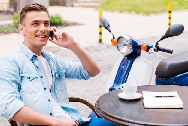 Gutes Gespräch mit Freund. Entspannter junger Mann, der am Handy spricht und lächelt, während er im Straßencafé mit Roller im Hintergrund sitzt