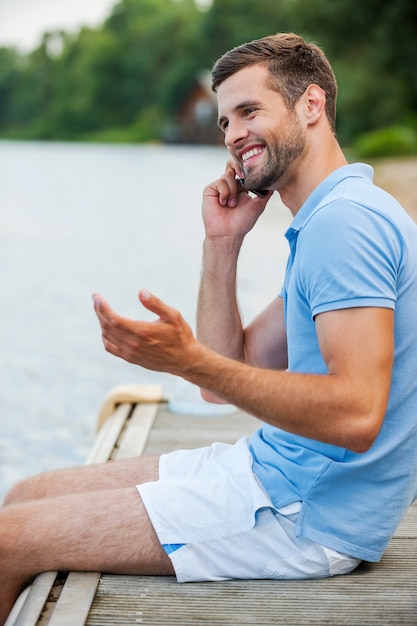 Gutes Gespräch mit Freund. Seitenansicht eines gutaussehenden jungen Mannes, der mit dem Handy spricht und lächelt, während er am Flussufer sitzt