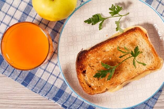 Gutes und leckeres Essen und Getränke zum Frühstück. Toast mit Butter und Käse auf Teller, Apfel und Glas Orangensaft auf Küchenserviette. Ansicht von oben.