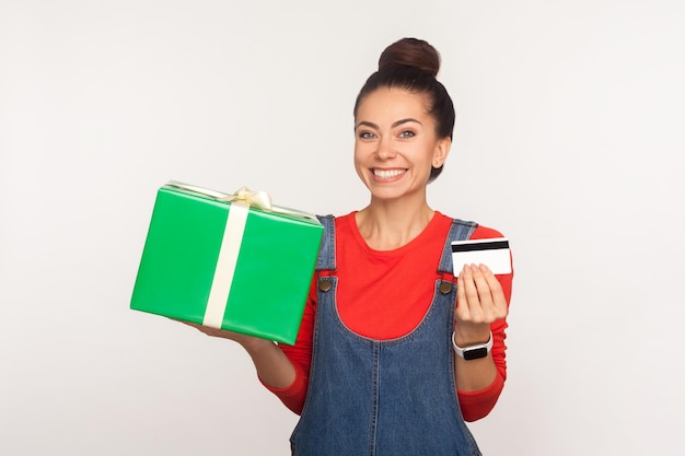 Guthaben für den Kauf von Weihnachtsgeschenken. Porträt eines stilvollen, fröhlichen Mädchens mit Haarknoten in Denim-Overalls, das eine Geschenkbox und eine Bankkarte hält und das Einkaufen mit Cashback, Bonus genießt. Indoor-Studioaufnahme isoliert