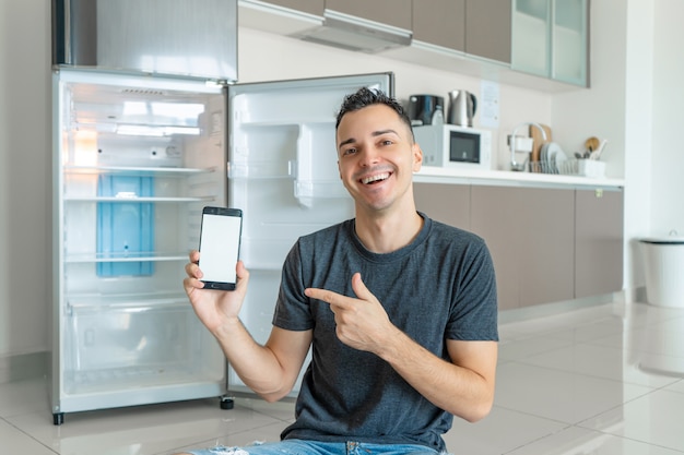 Guy bestellt Essen mit einem Smartphone in der Nähe von leeren Kühlschrank