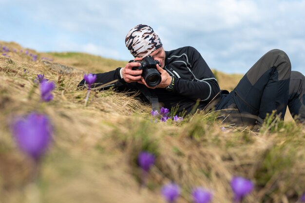 Guy Fotograf in einem Touristenoutfit liegt auf einer Lichtung und fotografiert Krokusblumen