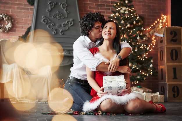 Guy gibt einen Kuss für seine Frau. Schöne Paare, die neues Jahr im verzierten Raum mit Weihnachtsbaum und Kamin hinten feiern