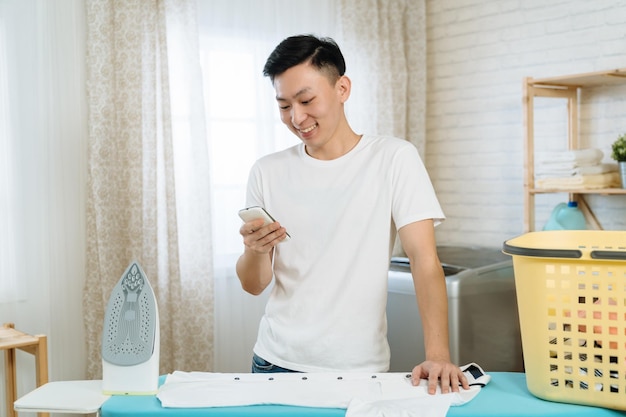 Guy sendet eine Nachricht auf dem Handy, während er zu Hause Wäsche an Bord bügelt. hausarbeit und technologiekonzept. lächelnder asiatischer chinesischer mann, der smartphone-bildschirm anschaut und online mit freunden chattet.