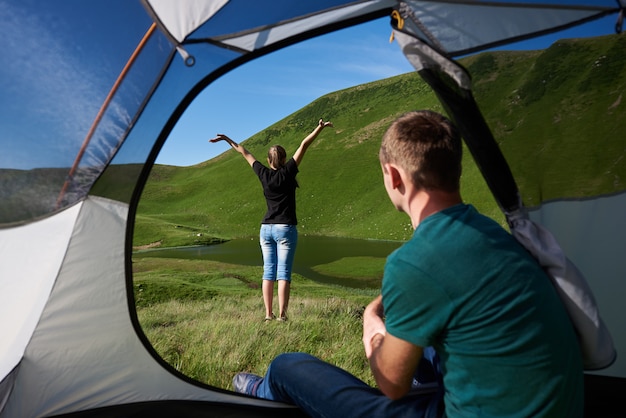 Foto guy sitzt in einem zelt und seine freundin genießt den blick auf einen klaren see am fuße eines mächtigen grünen berges unter blauem himmel. blick aus einem zelt