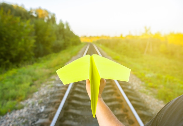 Guy sostiene un avión de papel amarillo hecho a mano. Foto del concepto de libertad con vías férreas. Motivación de estilo de vida de viaje. Industria del transporte ferroviario.