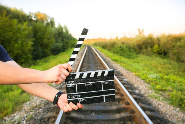 Guy sostiene claqueta negra en las manos. El hombre está dirigiendo y filmando una película de cine amateur. Senderos ferroviarios en el fondo, concepto de viaje.