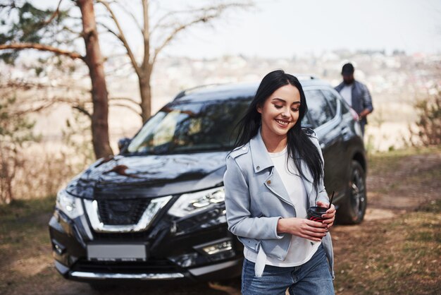 Guy toma equipo turístico de la parte trasera del automóvil. Pareja ha llegado al bosque en su nuevo coche negro