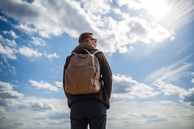 Guy turista autónomo con una mochila sobre los hombros contra un cielo azul con nubes blancas, día soleado