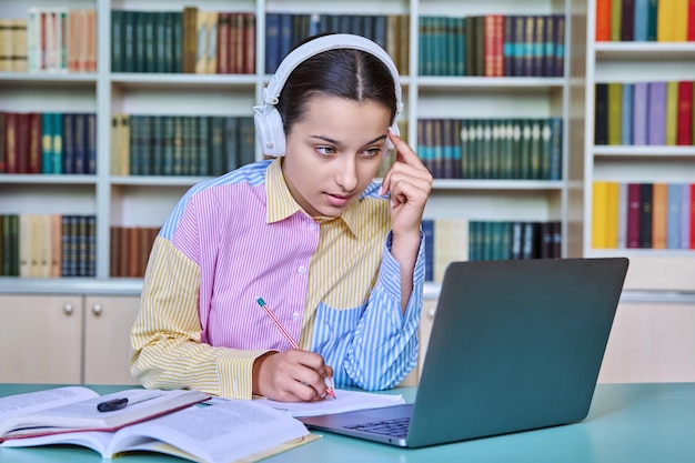 Gymnasiastin mit Kopfhörern studiert in der Schulbibliothek mit Büchern und Laptop