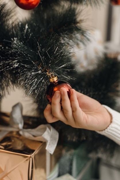 Una h en un suéter de punto blanco cuelga una bola roja de Navidad en la rama de un árbol de Navidad artificial. Decoración de árbol de Navidad con juguetes.